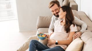 Cheerful young pregnant couple relaxing on a couch together, woman having healthy breakfast Cheerful young pregnant couple relaxing on a couch together, woman having healthy breakfast